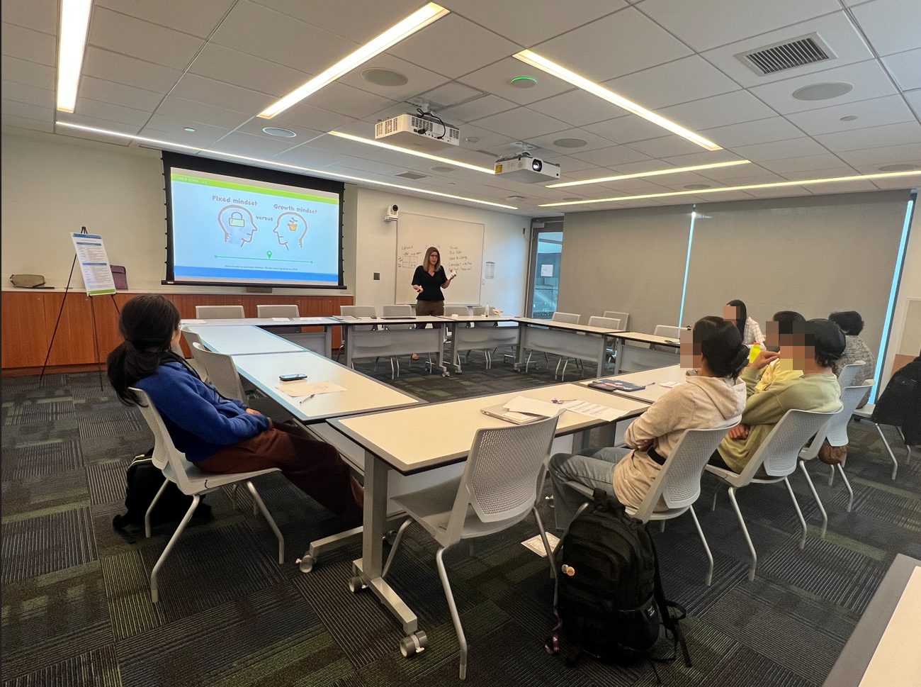 Image of conference room with tables set up in a square. Eleni is standing in the front of the room presenting.;